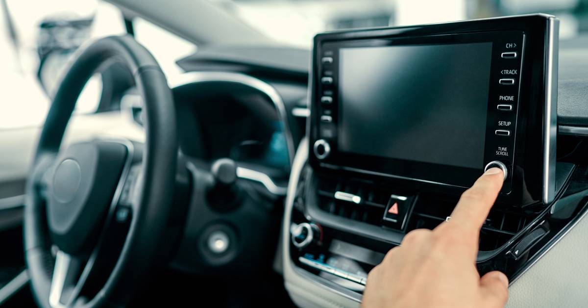 A person in the passenger seat pushing a dial on the car stereo. The stereo has a built-in touchscreen.